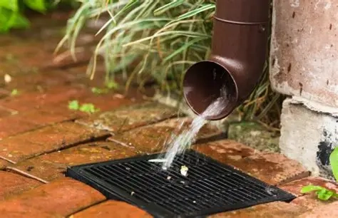 Rainwater pouring from brown downspout onto black grate in brick patio