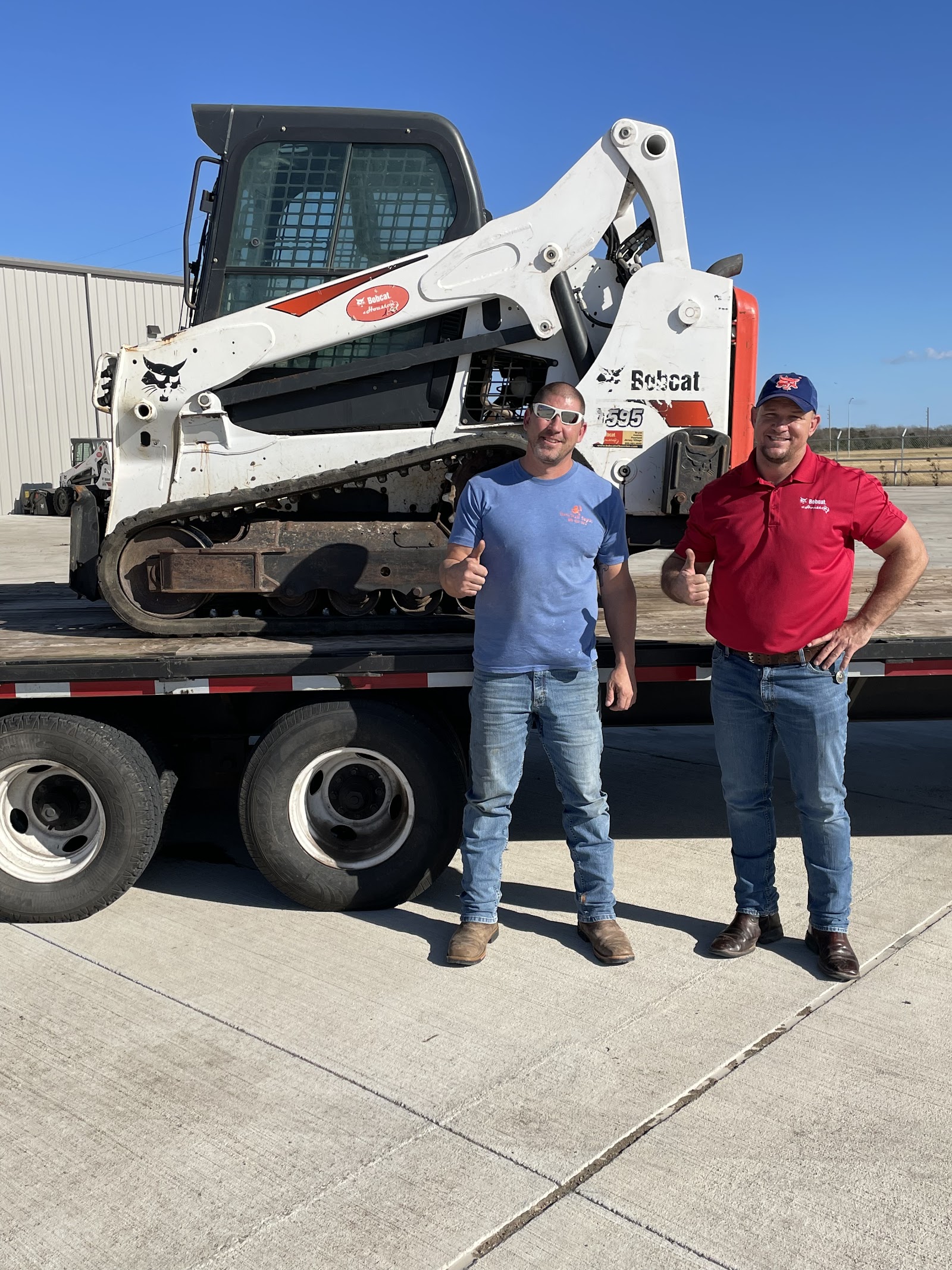 Two men posing in front of Bobcat skid steer loader on flatbed truck