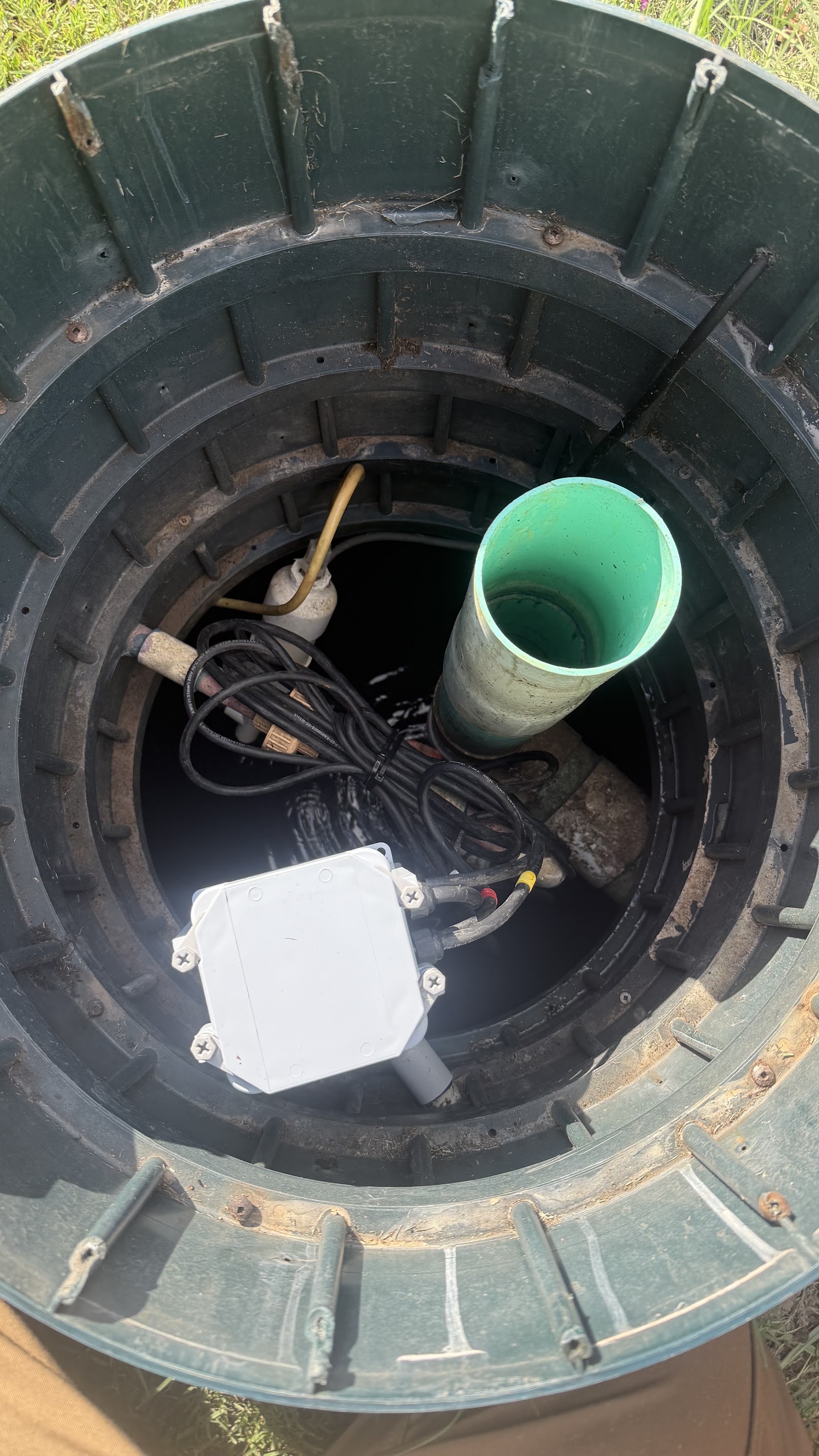 Overhead view inside a septic tank riser showing the pump, wiring, control box and green PVC outlet pipe