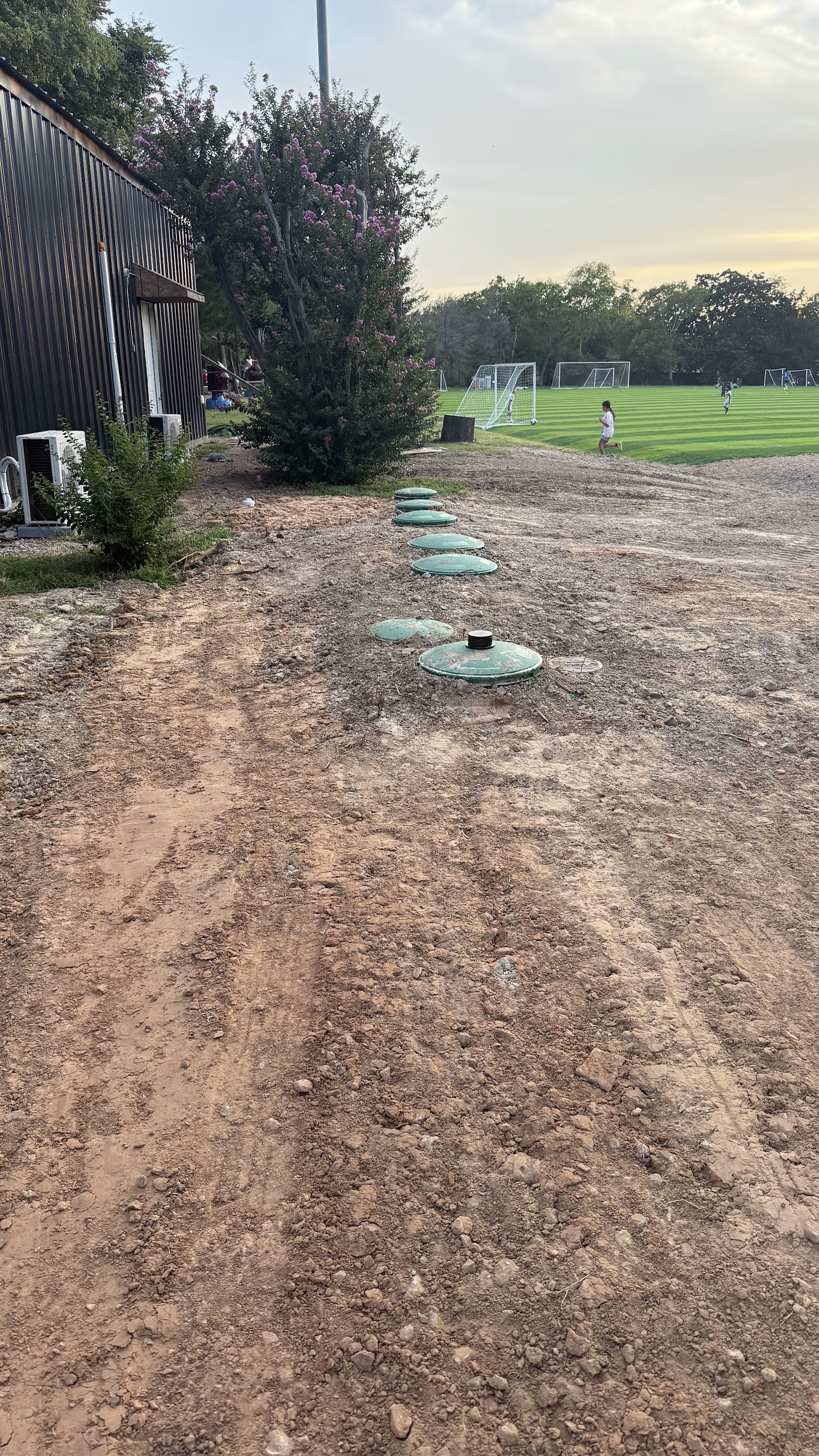 Row of green septic tank maintenance lids installed in dirt near a metal building and soccer field